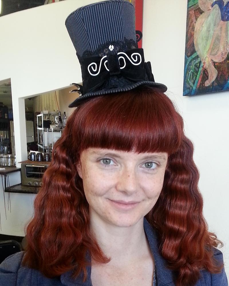 Copper-red hair with blunt fringe and crimped waves, styled with a mini top hat