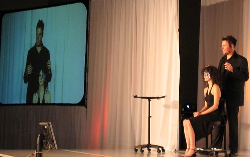 Christopher on a large stage beside a seated model, his work projected on a giant screen