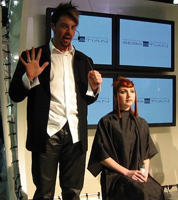 Christopher presenting in front of a wall of Sebastian monitors beside a model with a red bob