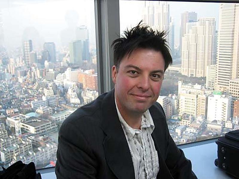 Christopher seated by a high-rise window with a sprawling city skyline behind him